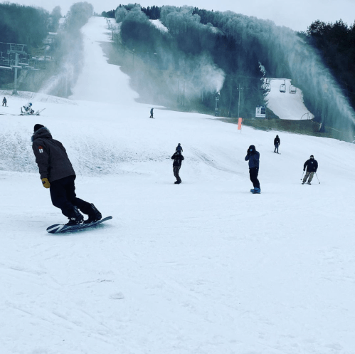 A snowy ski slope with several people skiing and snowboarding, surrounded by snow-making machines.
