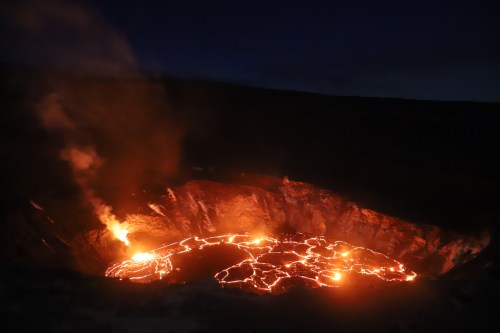 Lava flows in a volcanic crater at night, with smoke rising against a dark sky.