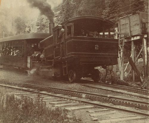 A vintage steam locomotive with a passenger car, traveling along tracks in a mountainous area.
