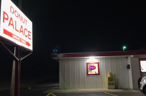 A brightly lit donut shop at night with a "Donut Palace" sign and an "Open" neon sign in the window.