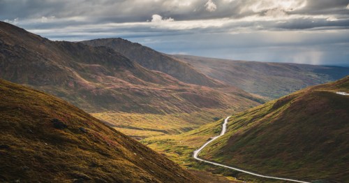 A winding road cuts through a lush, mountainous landscape under a dramatic sky with scattered clouds.
