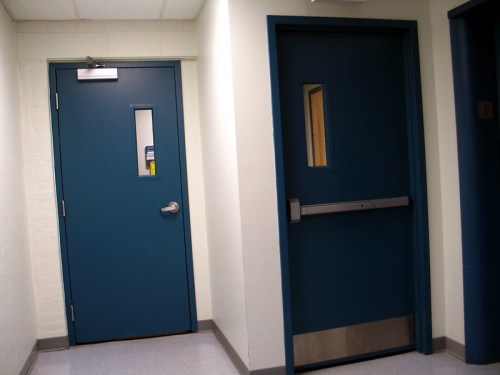 Two blue doors in a hallway, one with a small window and a handle, the other with a push bar and no window.