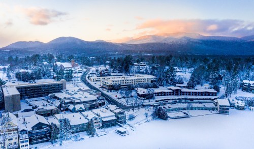 Aerial view of a snowy landscape with mountains, a lake, and a cluster of buildings in a winter setting.