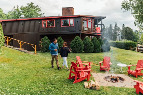 A couple walks on a grassy lawn near a fire pit, surrounded by red chairs and a cozy cabin in the background.