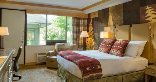 Cozy hotel room with a king bed, decorative pillows, armchair, and large window overlooking greenery.
