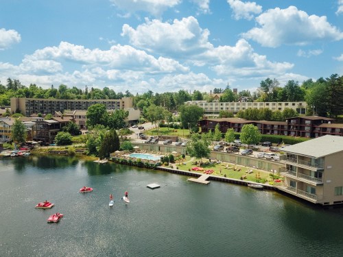 Aerial view of a lakeside resort with paddle boats, green lawns, and buildings under a blue sky with fluffy clouds.