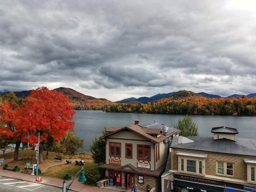 A scenic view of a lake surrounded by autumn foliage and mountains under a cloudy sky, with buildings in the foreground.