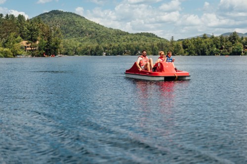 A family enjoys a sunny day on a red paddle boat in a serene lake surrounded by green mountains.