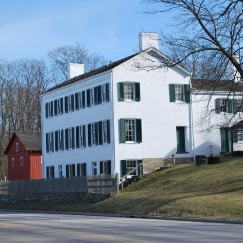 A white two-story house with green shutters, set against a backdrop of trees and a red barn in the distance.