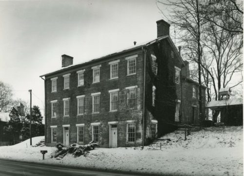 Historic brick building covered in snow, featuring multiple windows and a sloped yard with trees in the background.