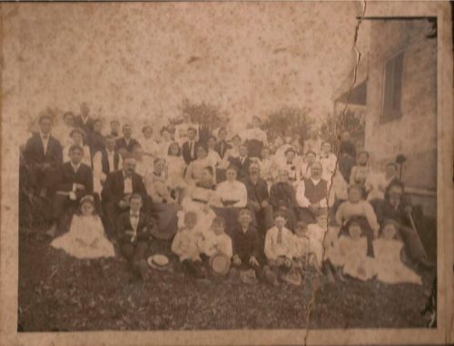 A large group of people, dressed in vintage clothing, poses together outdoors in a sepia-toned photograph.