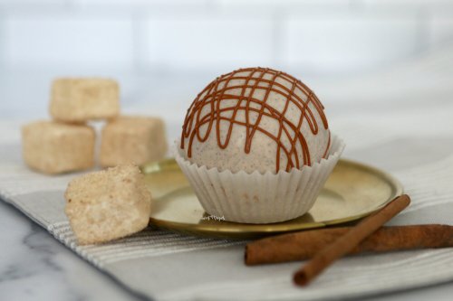 A decorative dessert ball drizzled with caramel, placed on a gold plate, with sugar cubes and a cinnamon stick nearby.