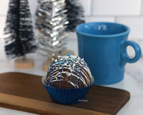 A chocolate truffle with blue and white drizzle on a wooden board, with a blue mug and decorative trees in the background.