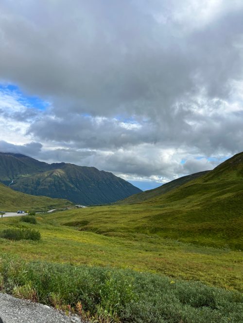 A scenic view of green hills and mountains under a cloudy sky, with a winding road in the distance.