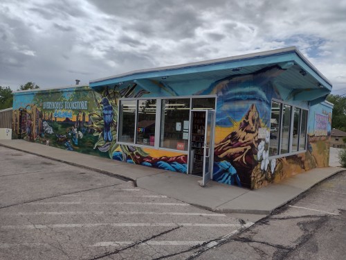 A colorful mural decorates the exterior of a bookstore, featuring mountains and flowers, with large windows and an open sign.