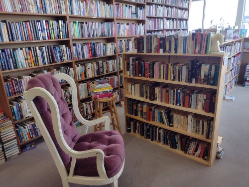A cozy reading nook with a plush chair beside a wooden bookshelf filled with books in a well-lit bookstore.