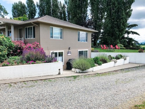 A house with a garden, surrounded by flowers, and a gravel driveway leading to a patio with red chairs.