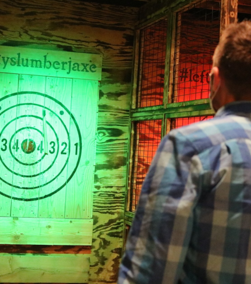 A person stands in front of a brightly lit axe-throwing target with a wooden backdrop.