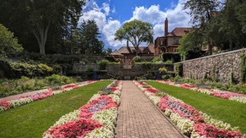 A scenic garden path lined with colorful flowers, leading to a stone wall and a large house under a blue sky.