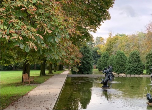 A serene park scene featuring a pond, sculptures, and trees with autumn foliage, alongside a walking path and bench.