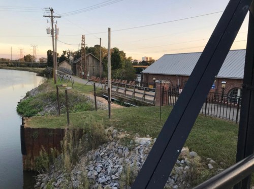 A calm riverbank with a gravel path, old buildings, and power lines in the background during sunset.