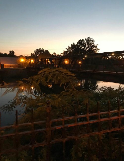A tranquil scene at dusk featuring a bridge over a calm waterway, surrounded by trees and soft lighting.