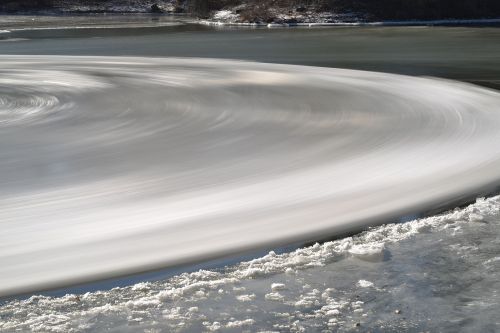 A swirling pattern of ice and water on a river, with a snowy bank in the foreground.