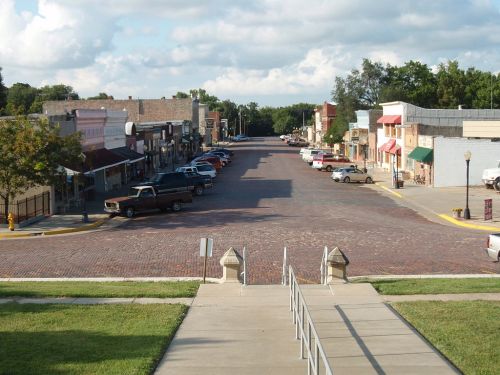 A view of a quiet, brick-paved street lined with shops and parked cars under a partly cloudy sky.