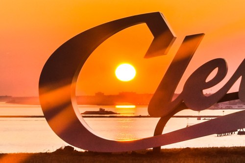 A sunset behind a large, stylized "C" sign, reflecting on calm waters with a warm orange sky.