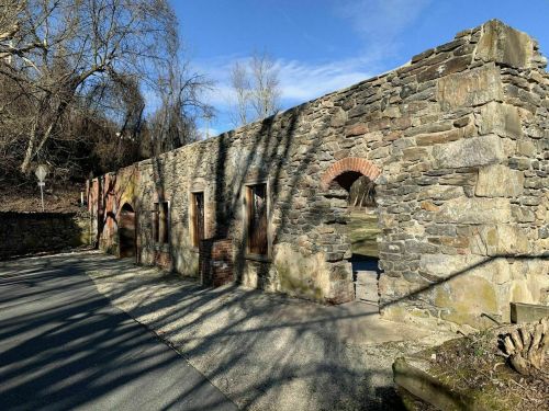 Ruins of a stone building with arched windows, surrounded by trees and a clear blue sky. Shadows cast on the ground.