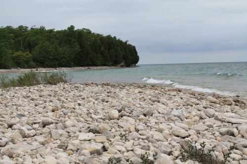 A rocky shoreline with gentle waves lapping at the stones, surrounded by green trees under a cloudy sky.