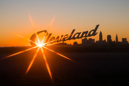 Sunset over Cleveland skyline with a large "Cleveland" sign in the foreground, creating a starburst effect.