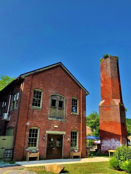 Historic red brick building with large windows and a tall chimney, set against a clear blue sky. Greenery on the chimney.