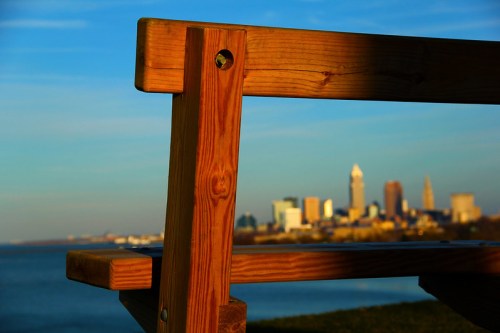 A wooden bench in the foreground with a city skyline and water in the background under a blue sky.
