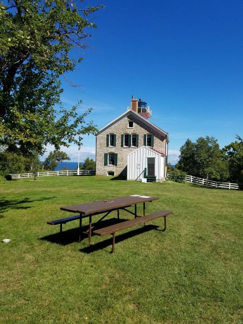 A historic building with green shutters, surrounded by grass and trees, features a picnic table in the foreground.