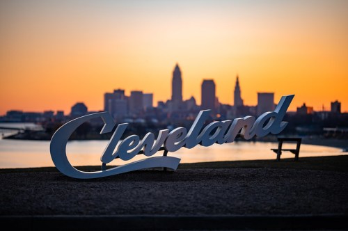 Cleveland sign in foreground with city skyline and sunset in the background, reflecting on the water.