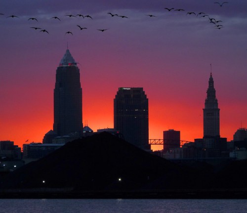 Silhouette of Cleveland skyline at sunset, with birds flying and a dark mound in the foreground.