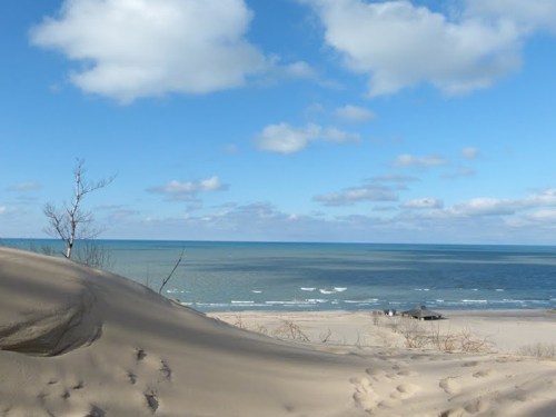 A sandy dune overlooks a calm lake under a blue sky with scattered clouds.