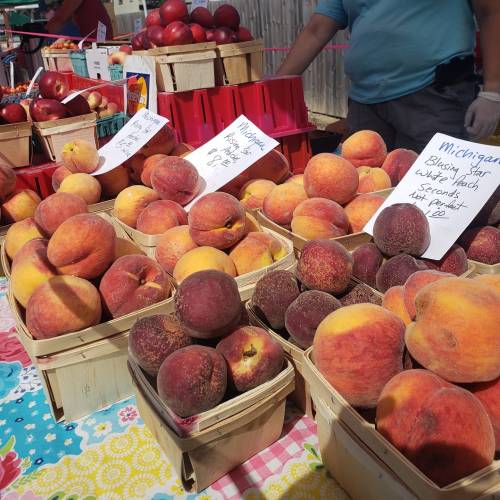 A vibrant farmers market display featuring baskets of fresh peaches and apples, with price signs visible.