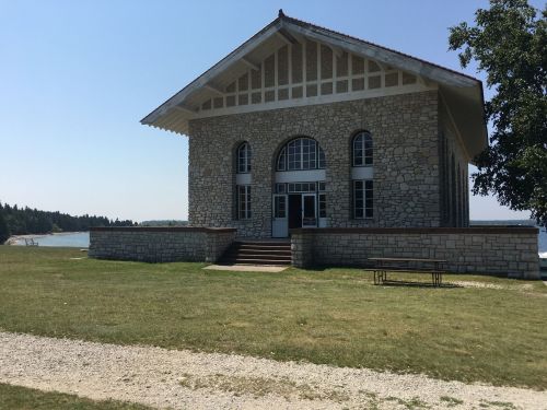 A large stone building with a gabled roof, set on a grassy area near a lake under a clear blue sky.