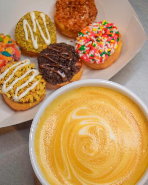A creamy latte with a swirl design, surrounded by colorful donuts in a box.