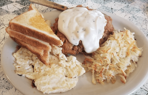 A plate of fried chicken topped with white gravy, served with toast, scrambled eggs, and hash browns.