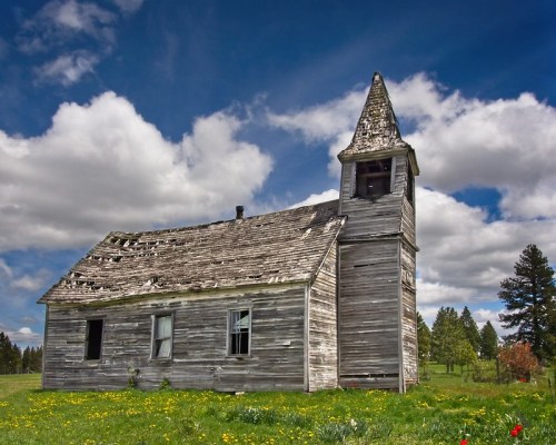 An old, weathered wooden church with a tall steeple, surrounded by green grass and wildflowers under a blue sky.