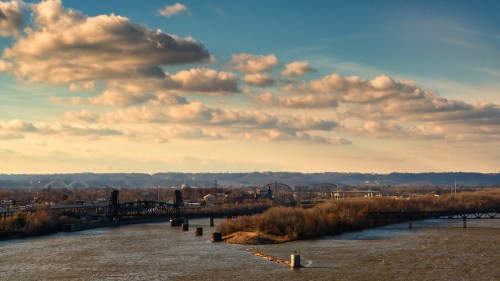 A scenic view of a river with bridges and clouds in the sky, surrounded by hills and trees.