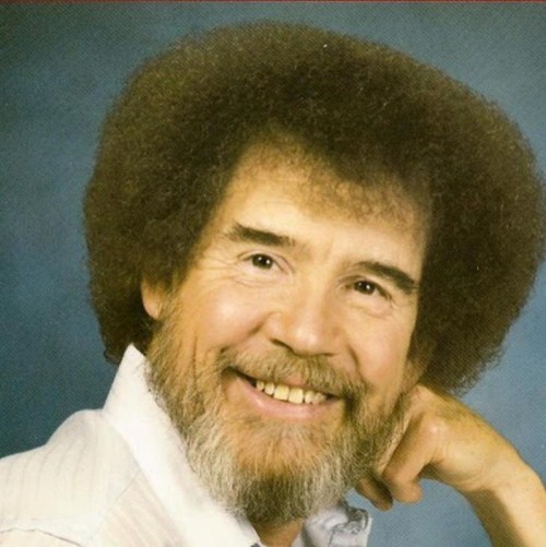 A smiling man with an afro hairstyle and a beard, wearing a white shirt, poses against a blue background.
