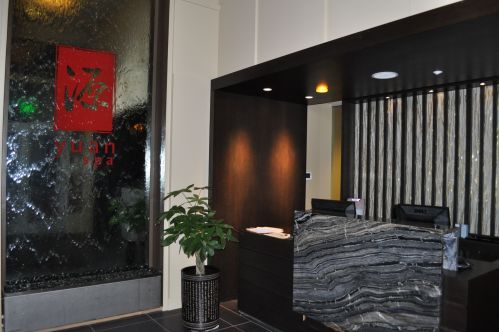Interior of a spa reception area featuring a water wall, a dark stone desk, and a potted plant. Red logo visible.