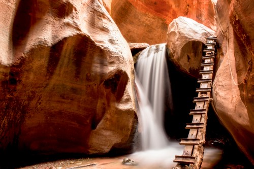 A serene waterfall cascades down rocky cliffs, with a wooden ladder leading up the side in a narrow canyon.