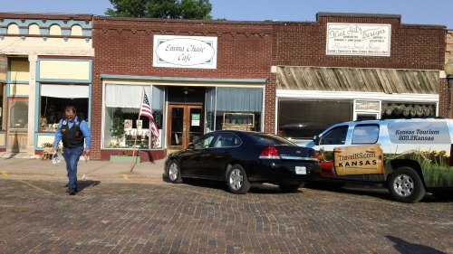 A man walks past a café and a tourism vehicle parked on a brick street, with an American flag displayed.