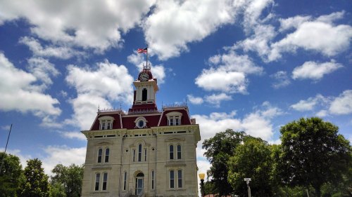Historic building with a clock tower, American flag, and a blue sky filled with clouds. Green trees in the foreground.