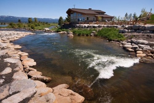 A serene river flows past a stone-lined bank, with a modern building and green landscape in the background under a clear blue sky.
